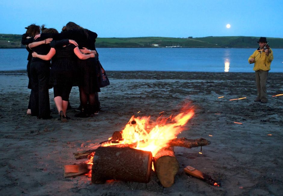 I am standing with a group of dancers. There is a fire burning on a beach, and a full moon. A fiddle player stands against in front of the sea.