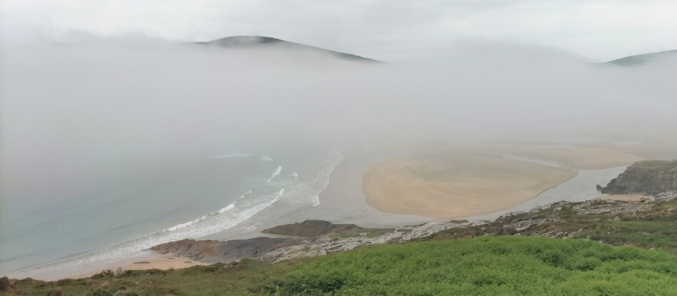 Thick fog covers the view of a mountain. Photo also shows the sea, a beach, and grass in the foreground.