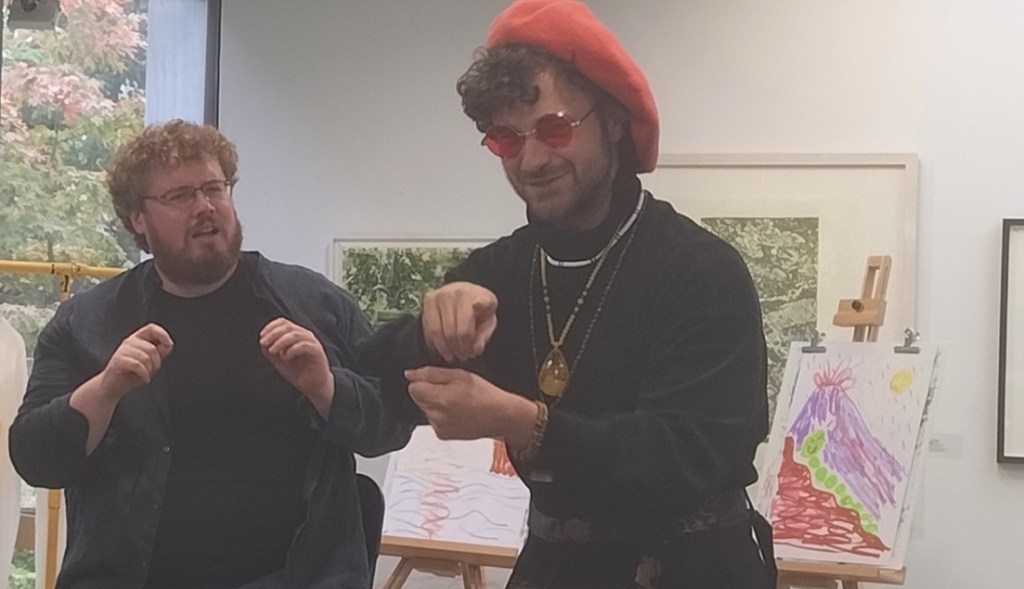 Two actors in an art gallery performing a play. One wears an orange beret and is standing up. The other is seated.